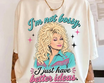 May include: An ivory short-sleeve t-shirt featuring a graphic illustration of a blonde woman with voluminous hair wearing a teal shirt with fringe. The text reads I'm not bossy, I just have better ideas in teal and pink script and block lettering.