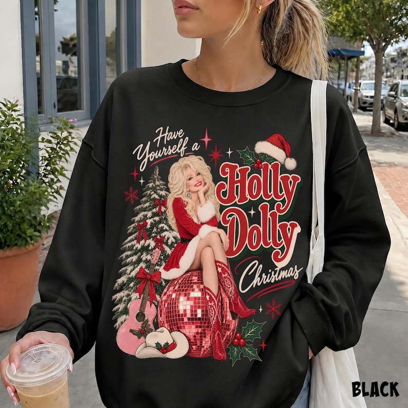 May include: A black long-sleeve sweatshirt featuring a graphic print of a blonde woman in a red outfit sitting on a disco ball. The text reads Have Yourself a Holly Dolly Christmas. A Christmas tree, a guitar, and a cowboy hat are also depicted.