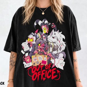Op de afbeelding: Een zwart t-shirt met een grafische print van vier geanimeerde personages uit Helluva Boss. De personages zijn omringd door reisartikelen zoals een koffer, paspoort en kaart. De tekst "OUT OF OFFICE" staat onderaan in grote rode letters gedrukt.