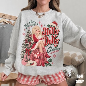 May include: An ash gray crewneck sweatshirt featuring a graphic print of a blonde woman in a red dress and cowboy boots sitting on a disco ball. The text reads Have Yourself a Holly Dolly Christmas, accompanied by a pine tree and guitar.