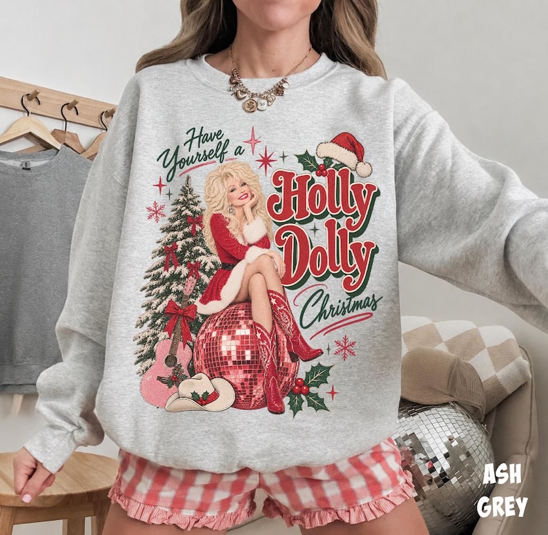 May include: An ash gray crewneck sweatshirt featuring a graphic print of a blonde woman in a red dress and cowboy boots sitting on a disco ball. The text reads Have Yourself a Holly Dolly Christmas, accompanied by a pine tree and guitar.