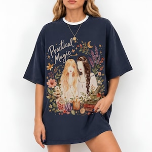 May include: A dark navy blue short-sleeve t-shirt featuring a graphic print with the text "Practical Magic" above two figures surrounded by flowers, a crescent moon, a jar labeled "Rose Petals," a candle, and a stack of books labeled "Spells."