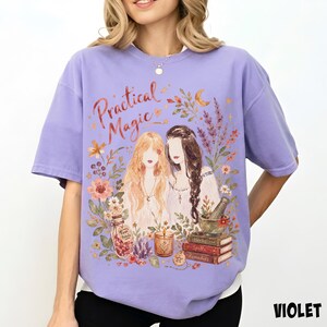May include: A light purple t-shirt featuring a graphic illustration of two figures with long hair surrounded by flowers, a crescent moon, a jar labeled Rose Petals, a candle, a mortar and pestle, and a stack of books labeled Herbs, Spells, and Remedies.