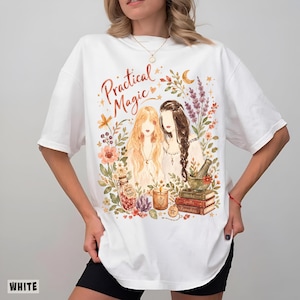 May include: A white short-sleeve t-shirt featuring a watercolor-style graphic with the text "Practical Magic". The design includes two figures surrounded by flowers, a bottle labeled "Rose Petals", a candle, a mortar and pestle, and stacked books.