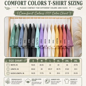 以下が含まれることがあります： Comfort Colors 1717 Tシャツのカラーチャート。木製のラックに、Light Green、Moss、Blue Jean、Gray、Black、Pepper、Chalky Mint、Violet、Orchid、White、Ivory、Mustard、Yam、Cumin、Berryとラベル付けされた15色のTシャツが掛けられています。