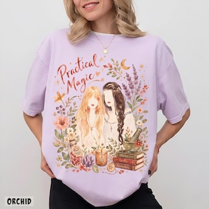 May include: A light purple t-shirt featuring a graphic print with the text "Practical Magic" above two figures surrounded by flowers, a crescent moon, a jar labeled "Rose Petals," a candle, a mortar and pestle, and a stack of books labeled "Herbs," "Spells," and "Remedies."