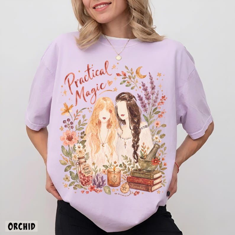 May include: A light purple t-shirt featuring a graphic print with the text "Practical Magic" above two figures surrounded by flowers, a crescent moon, a jar labeled "Rose Petals," a candle, a mortar and pestle, and a stack of books labeled "Herbs," "Spells," and "Remedies."