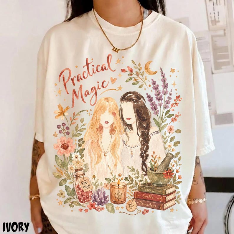 May include: An ivory t-shirt featuring a graphic illustration of two figures with long hair surrounded by flowers, lavender, a bottle labeled Rose Petals, a candle, and a stack of books labeled Herbs, Spells, and Remedies with the text Practical Magic.