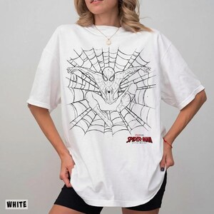 以下が含まれることがあります： 蜘蛛の巣の中央にスパイダーマンが描かれた黒い線画のグラフィックが特徴の白い半袖Tシャツ。衣服の前面、イラストの下には、赤と黒の文字でマーベル・スタジオのスパイダーマンのロゴがプリントされています。