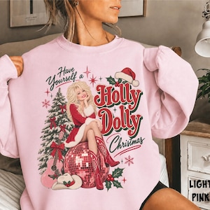 May include: A light pink crewneck sweatshirt featuring a graphic print of a blonde woman in a red dress sitting on a disco ball. The text reads Have Yourself a Holly Dolly Christmas. Includes a Christmas tree, guitar, and cowboy boots.