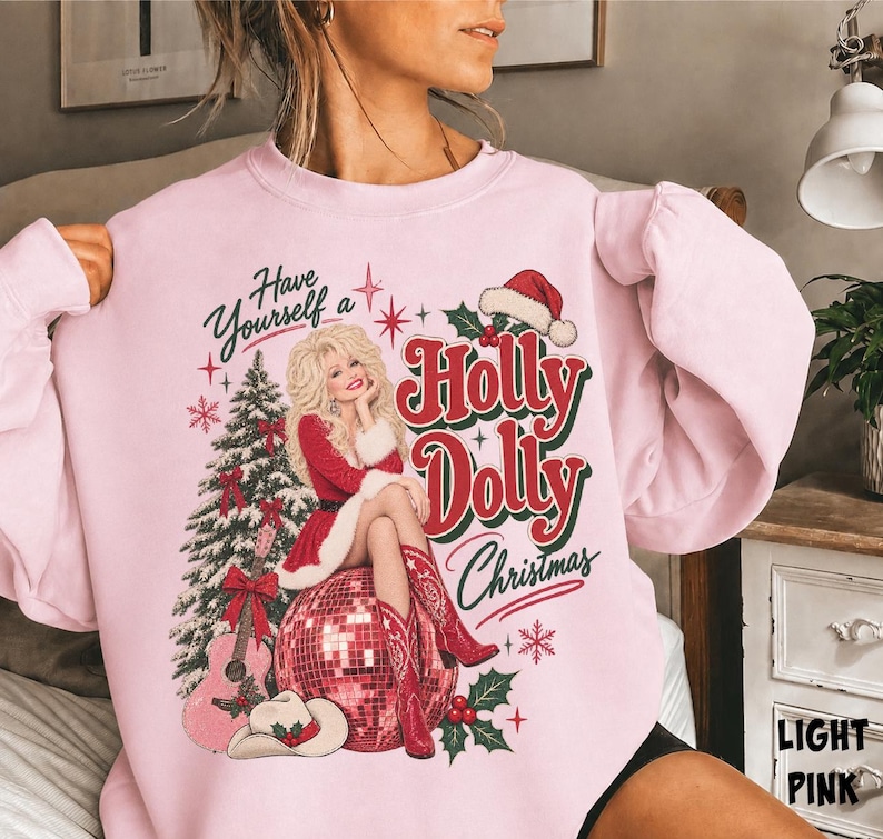 May include: A light pink crewneck sweatshirt featuring a graphic print of a blonde woman in a red dress sitting on a disco ball. The text reads Have Yourself a Holly Dolly Christmas. Includes a Christmas tree, guitar, and cowboy boots.