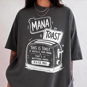 May include: A charcoal gray t-shirt featuring a white graphic of a toaster with a slice of bread popping out. The text reads: MANA TOAST, THIS IS TOAST. IT REFILLS YOUR MANA. THAT'S IT. NOTHING MORE. F#!$ YOU. The design includes small star and heart accents.