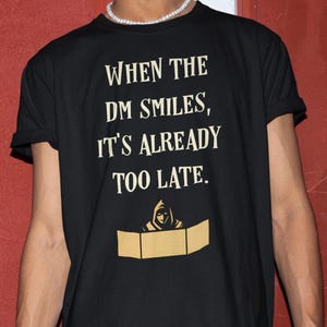 May include: A black short-sleeved t-shirt featuring the cream-colored text "WHEN THE DM SMILES, IT'S ALREADY TOO LATE." Below the text is a graphic of a hooded figure positioned behind a three-paneled screen, rendered in a matching cream color.