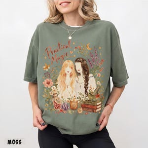 May include: A moss green crewneck t-shirt featuring a graphic print of two figures surrounded by flowers, a crescent moon, a bottle labeled 'First Petals', a candle, and a stack of books labeled 'Herbs', 'Spells', and 'Remedies' with the text 'Practical Magic'.