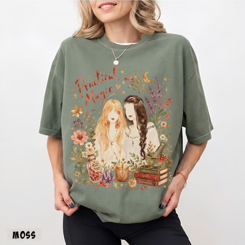 May include: A moss green crewneck t-shirt featuring a graphic print of two figures surrounded by flowers, a crescent moon, a bottle labeled 'First Petals', a candle, and a stack of books labeled 'Herbs', 'Spells', and 'Remedies' with the text 'Practical Magic'.