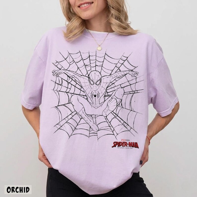 以下が含まれることがあります： 大きなクモの巣のデザインの中央にスパイダーマンの黒い線画が描かれた、ライトパープルの半袖Tシャツ。グラフィックの右下隅には、赤と黒のスパイダーマンのロゴとテキストが含まれています。