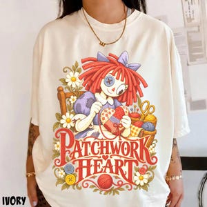 Peut inclure: Un t-shirt ivoire orné d'un motif représentant une poupée aux cheveux roux avec un œil en bouton, cousant un cœur en patchwork. Le dessin inclut des marguerites, de la laine, des ciseaux et le texte rouge "PATCHWORK HEART" avec des boutons.
