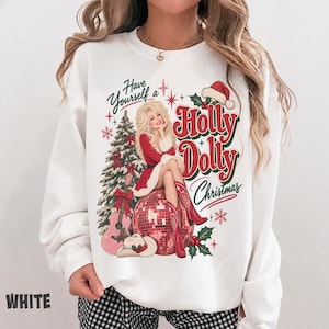 May include: A white crewneck sweatshirt featuring a graphic print of a blonde woman in a red dress sitting on a disco ball. The design includes a Christmas tree, a guitar, a cowboy hat, and the text Have Yourself a Holly Dolly Christmas.