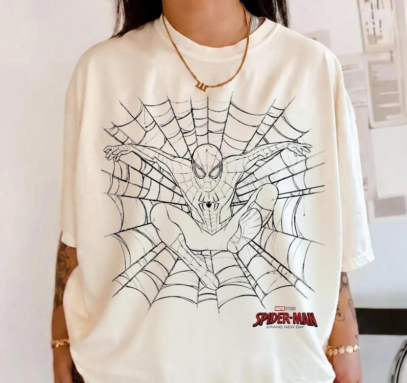 以下が含まれることがあります： オフホワイトの半袖Tシャツ。中央にクモの巣とスパイダーマンの黒い線画が描かれています。右下には赤と黒の「SPIDER-MAN BRAND NEW DAY」のロゴがあります。クルーネックでゆったりとしたフィット感のTシャツです。