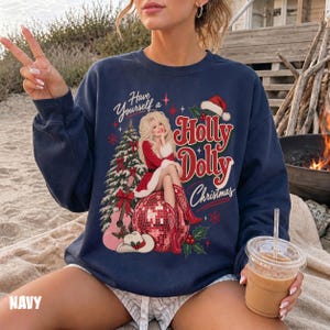May include: A navy blue crewneck sweatshirt featuring a graphic print of a blonde woman in a red dress sitting on a red disco ball. The text reads Have Yourself a Holly Dolly Christmas. A Christmas tree, guitar, and cowboy hat are included.