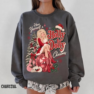 May include: A charcoal gray crewneck sweatshirt featuring a graphic print of a blonde figure in a red dress and cowboy boots sitting on a disco ball. The design includes a Christmas tree, a guitar, a cowboy hat, and the text Have Yourself a Holly Dolly Christmas.
