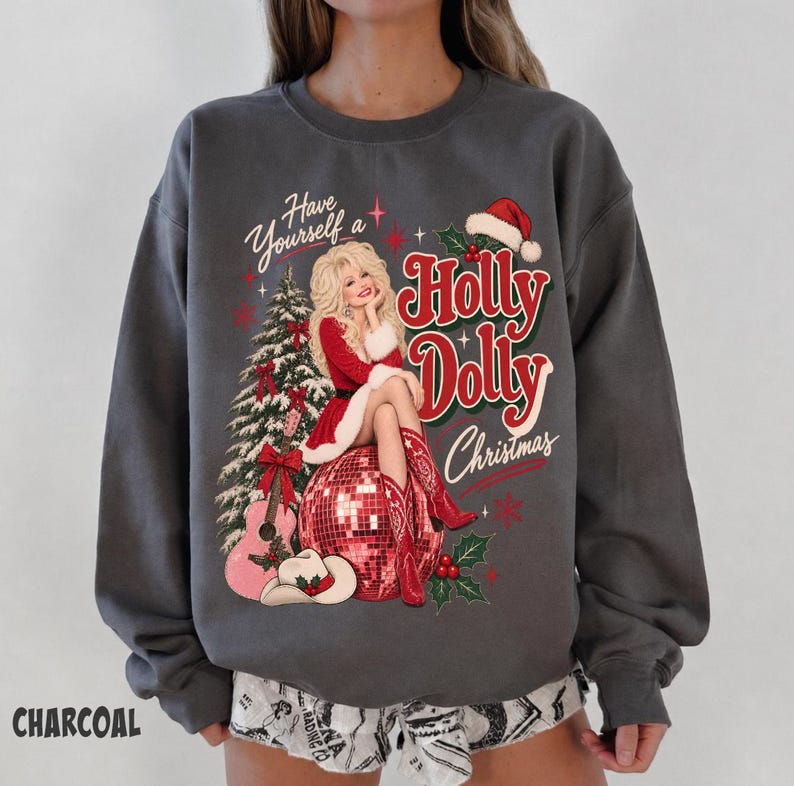 May include: A charcoal gray crewneck sweatshirt featuring a graphic print of a blonde figure in a red dress and cowboy boots sitting on a disco ball. The design includes a Christmas tree, a guitar, a cowboy hat, and the text Have Yourself a Holly Dolly Christmas.