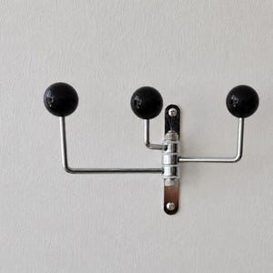 May include: A wall-mounted coat rack featuring a vertical chrome metal plate with three extending arms. Each arm ends in a smooth, round, black ball-shaped hook. The central mechanism allows the arms to pivot, and the unit is secured with two screws.