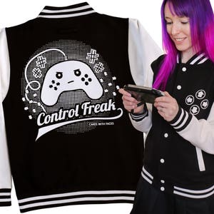 May include: A black and white varsity jacket with a white and black striped trim. The jacket has a white graphic of a video game controller with a face on it and the text "Control Freak" with the words "Cakes with Faces" underneath. A person is wearing the jacket and holding a video game controller.