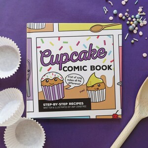 May include: A colorful cupcake comic book with step-by-step recipes. The cover features two cartoon cupcakes with icing and sprinkles. The book is on a purple surface with cupcake liners and a wooden spoon.