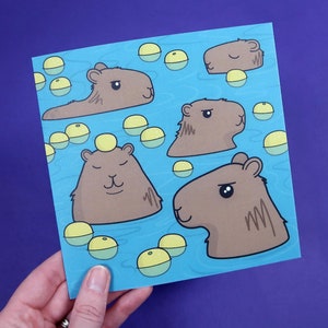 Puede incluir: Una tarjeta azul con una ilustración de dibujos animados de cinco capibaras flotando en una piscina de agua con círculos amarillos. Los capibaras son marrones y tienen ojos y narices negros.