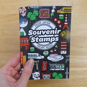 Puede incluir: Un folleto negro "Souvenir Stamps" de Japón, decorado con coloridas ilustraciones de la cultura japonesa. La portada muestra sushi, un tren, la Torre Eiffel y un gato de la suerte.