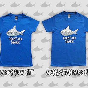 May include: Two blue t-shirts with a graphic of a cartoon shark with white frosting and sprinkles. The text on the shirts reads "Great Iced Shark". The shirt on the left is labeled "Ladies Slim Fit" and the shirt on the right is labeled "Mens Standard Fit".