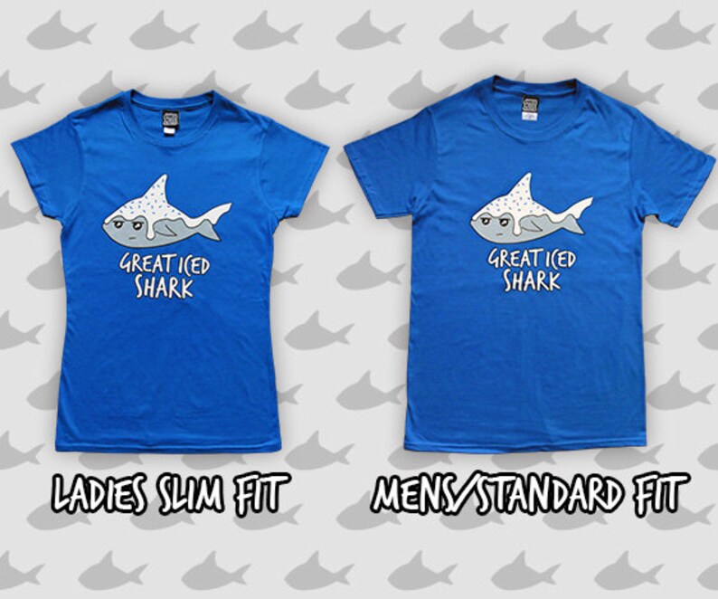 May include: Two blue t-shirts with a graphic of a cartoon shark with white frosting and sprinkles. The text on the shirts reads "Great Iced Shark". The shirt on the left is labeled "Ladies Slim Fit" and the shirt on the right is labeled "Mens Standard Fit".
