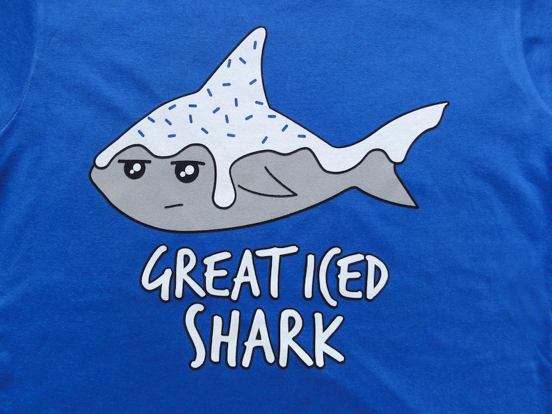 May include: A blue t-shirt with a cartoon illustration of a gray shark with white frosting and blue sprinkles. The text "Great Iced Shark" is printed below the shark.
