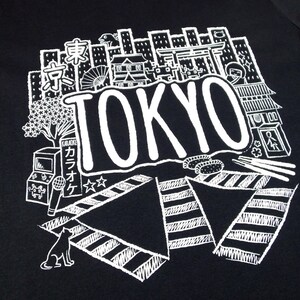 May include: Black t-shirt with a white graphic design of a cityscape with the word "TOKYO" in large letters. The design includes various Japanese cultural elements like cherry blossoms, a torii gate, a karaoke machine, and a cat.
