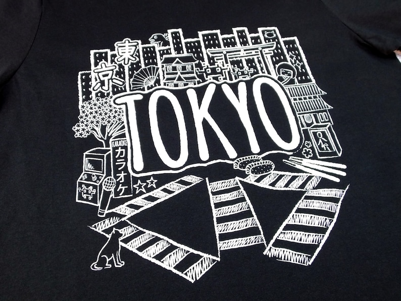 May include: Black t-shirt with a white graphic design of a cityscape with the word "TOKYO" in large letters. The design includes various Japanese cultural elements like cherry blossoms, a torii gate, a karaoke machine, and a cat.
