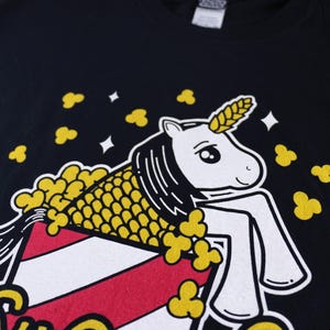 May include: A black t-shirt with a white unicorn wearing a yellow popcorn costume. The unicorn is sitting in a red and white striped popcorn bucket.