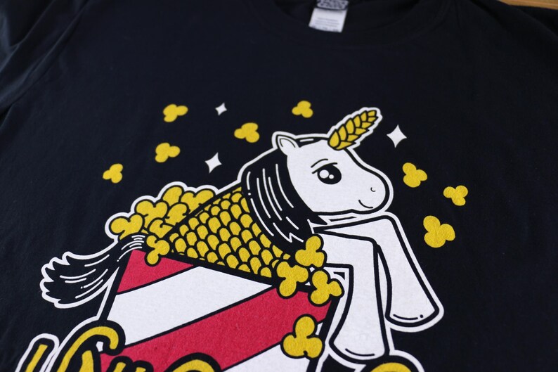 May include: A black t-shirt with a white unicorn wearing a yellow popcorn costume. The unicorn is sitting in a red and white striped popcorn bucket.