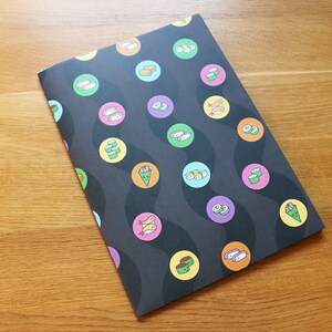 May include: A black notebook with a sushi pattern. The notebook has a wavy design with circles containing different types of sushi. The sushi is illustrated in orange, green, and yellow.