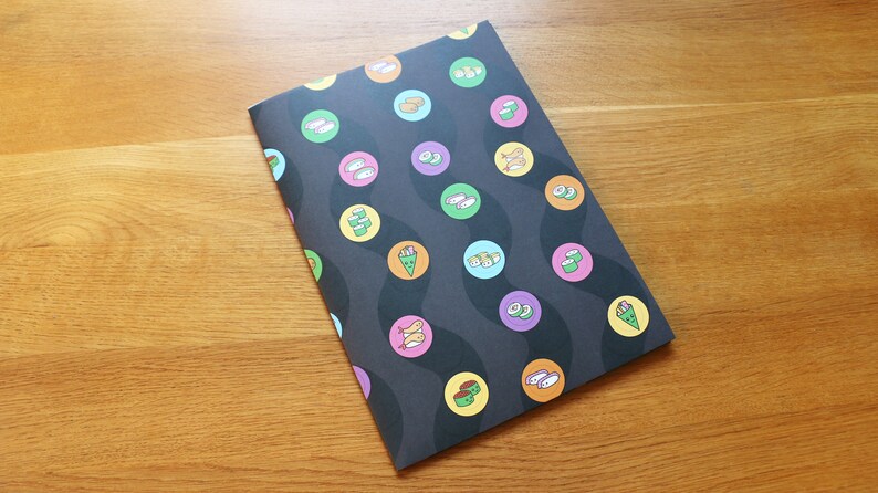 May include: A black notebook with a sushi pattern. The notebook has a wavy design with circles containing different types of sushi. The sushi is illustrated in orange, green, and yellow.