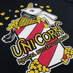 May include: A black t-shirt with a graphic of a unicorn inside a popcorn bucket. The text "Unicorn" is in yellow letters and the text "Magical Buttery Goodness" is in white letters.