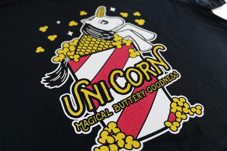 May include: A black t-shirt with a graphic of a unicorn inside a popcorn bucket. The text "Unicorn" is in yellow letters and the text "Magical Buttery Goodness" is in white letters.