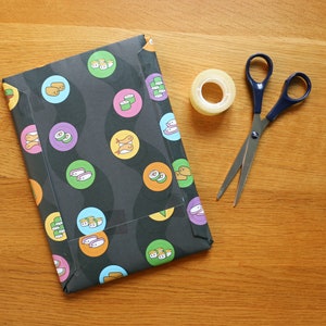 May include: Black and white patterned wrapping paper with sushi and fish designs. A roll of clear tape and a pair of silver scissors with blue handles are on a wooden surface.