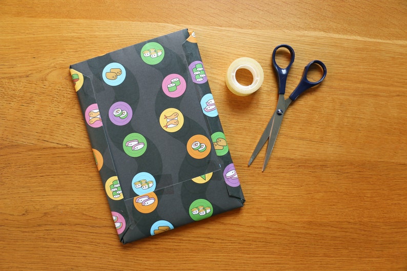 May include: Black and white patterned wrapping paper with sushi and fish designs. A roll of clear tape and a pair of silver scissors with blue handles are on a wooden surface.