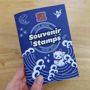 May include: A blue booklet titled Souvenir Stamps From Japan featuring a white graphic of a beckoning cat, stylized ocean waves, a fish, and an origami crane. The cover includes the text Cakes with Faces and a repeating geometric pattern.