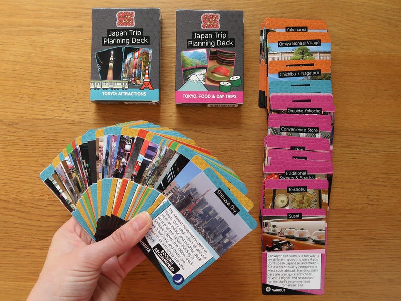 Op de afbeelding: Een set kleurrijke speelkaarten met afbeeldingen van Japanse bezienswaardigheden, eten en activiteiten. De kaarten zijn verdeeld in twee decks: "Tokyo Attracties" en "Tokyo: Eten & Dagtrips". Er is ook een stapel kaarten met verschillende Japanse locaties en activiteiten.