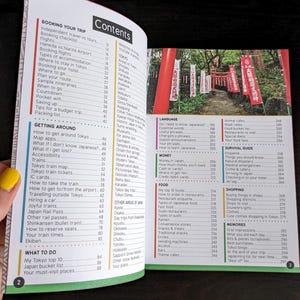 Pu&ograve; includere: Guida turistica aperta con un sommario e una sezione su Tokyo. La pagina di destra mostra una foto di un santuario giapponese con porte torii rosse e striscioni. Il libro &egrave; aperto su una superficie scura.