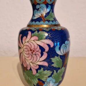 May include: A cloisonné vase featuring a deep blue background with a repeating cloud pattern. The vase is decorated with large pink and light blue chrysanthemum flowers and green leaves, all outlined in thin gold metal wire with a gold rim.