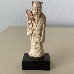 May include: A small, off-white carved figurine depicting a bearded figure in traditional robes holding a fan. The figure features intricate etched details and is mounted on a solid, square, black base, creating a contrast in colour and texture.