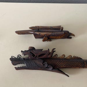 Op de afbeelding: Twee donkerbruine, uit hout gesneden objecten. Het bovenste stuk is een vogel met gedetailleerde vleugel- en veerpatronen. Het onderste stuk is een lange, horizontale drakenfiguur met een open bek, zichtbare tanden en ingewikkelde schubben.